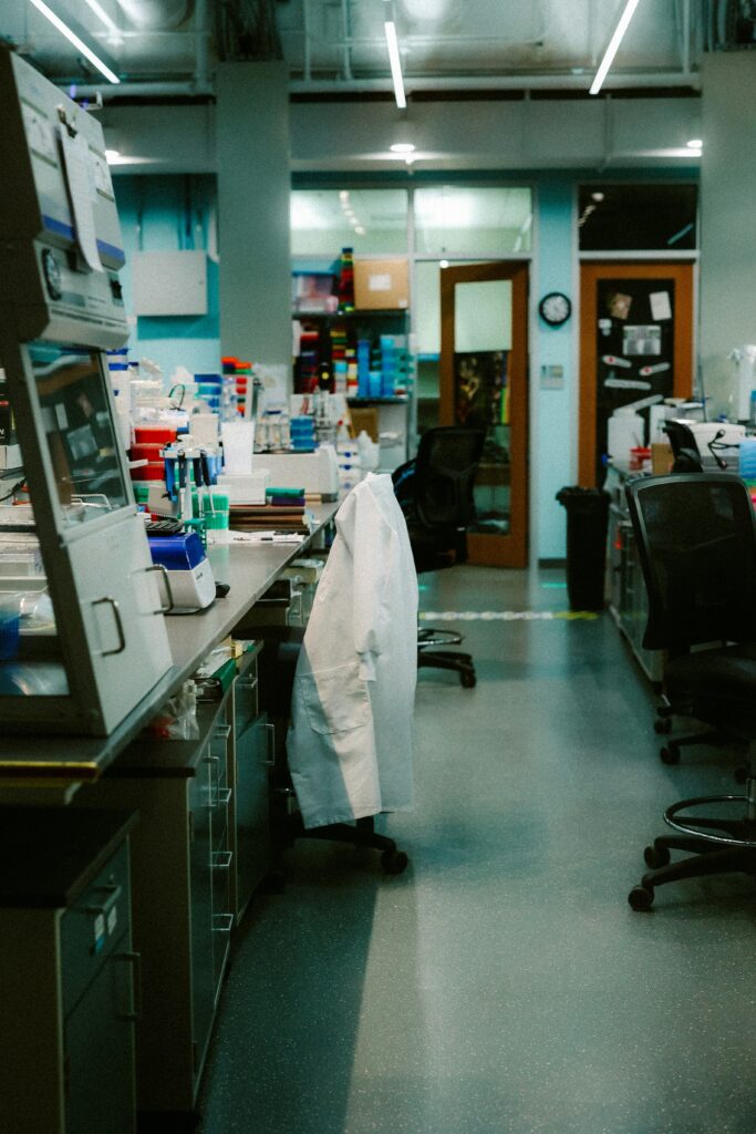 Busy laboratory workstation with benches and shelves; a white lab coat draped over a chair in the foreground.
