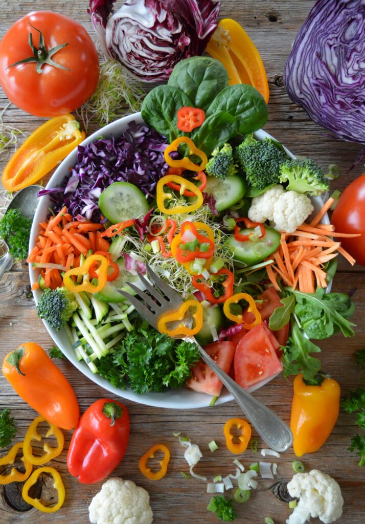 Colorful vegetable salad in a white bowl on a wooden table, featuring bell peppers, tomatoes, cucumber, broccoli, cauliflower, red cabbage, carrots, and greens.
